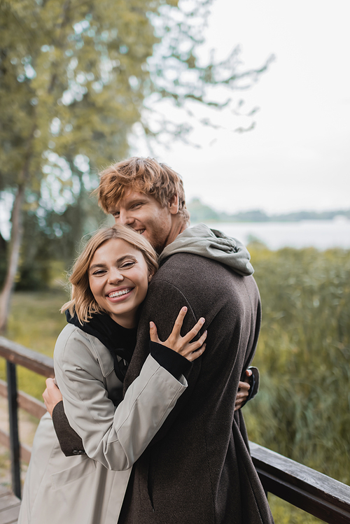 joyful young woman smiling and hugging redhead man during date on bridge near pond