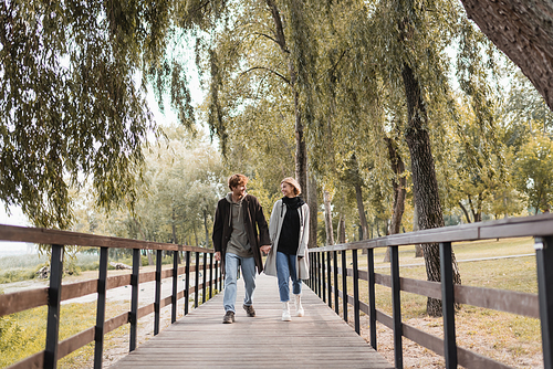 full length of redhead man and blonde woman in coats smiling while holding hands and walking on bridge