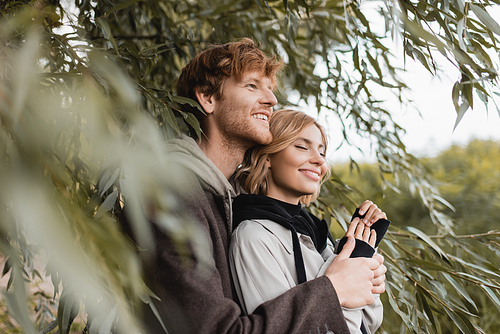 happy man hugging cheerful young woman near green leaves on blurred foreground