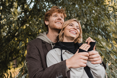 low angle view of happy man hugging cheerful young woman near green leaves on tree