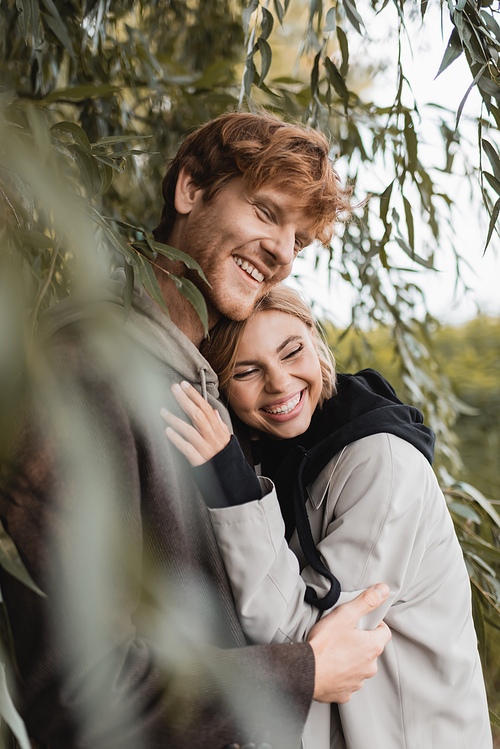 happy blonde woman hugging cheerful young man near green leaves on blurred foreground