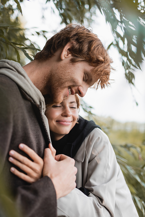 pleased blonde woman hugging joyful young man near green leaves on tree