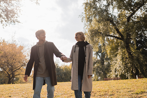 young and happy couple looking at each other while holding hands in autumnal park