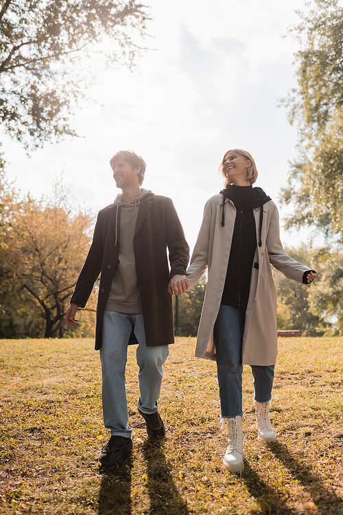 full length of young and happy couple holding hands while walking in autumnal park