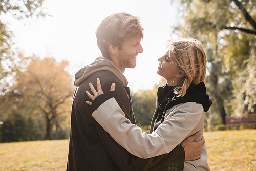 side view of young and happy couple smiling while hugging in autumnal park