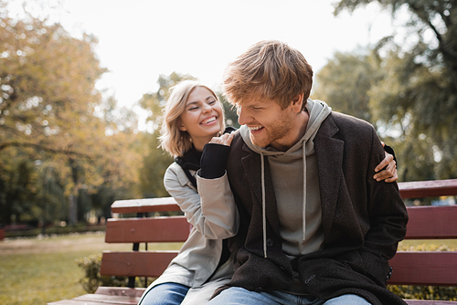 happy and blonde woman hugging redhead boyfriend while sitting on bench in park