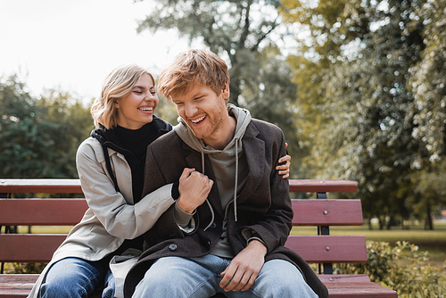 cheerful and blonde woman embracing redhead boyfriend while sitting on bench in park