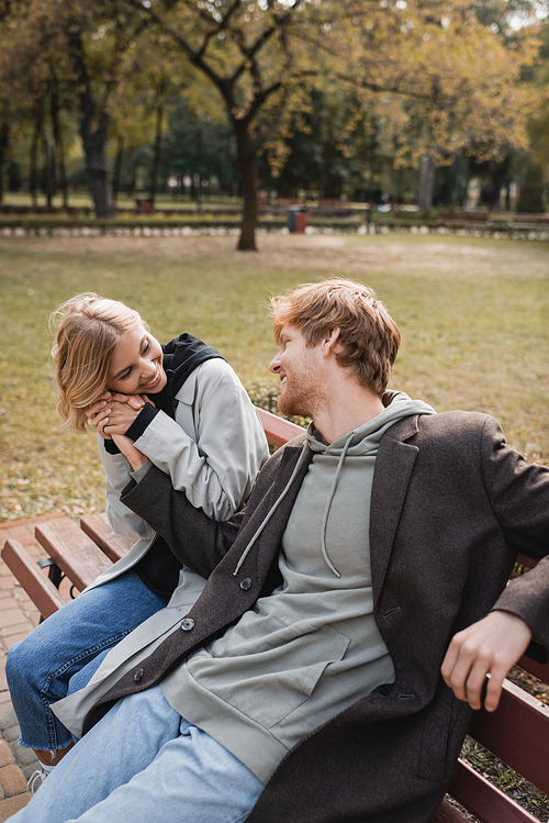 cheerful and blonde woman hugging hand of redhead boyfriend while sitting on bench in park