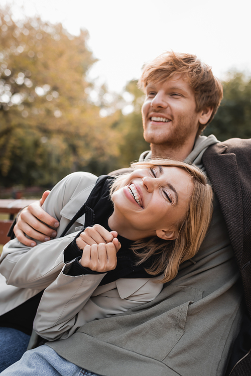 redhead man embracing happy blonde woman while smiling in autumnal park