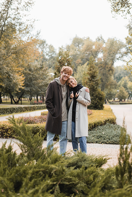 joyful and young man in coat hugging blonde girlfriend while standing around plants in park