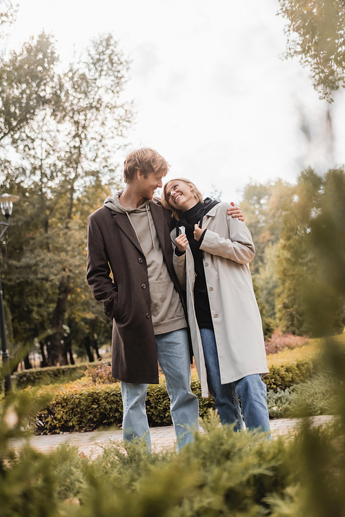 happy and young man in coat hugging blonde girlfriend while standing around plants in park