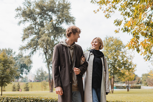 young and positive couple in coats looking at each other while walking in autumnal park during date