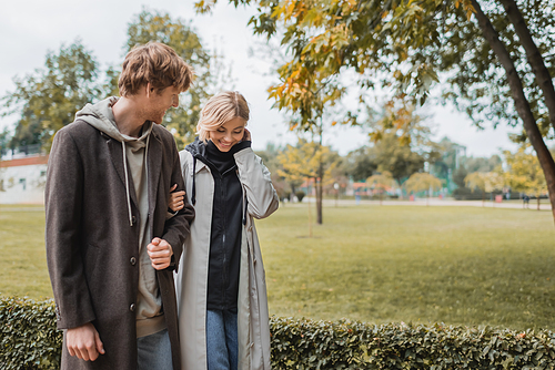 young and positive man in coat walking with blonde girlfriend in autumnal park during date