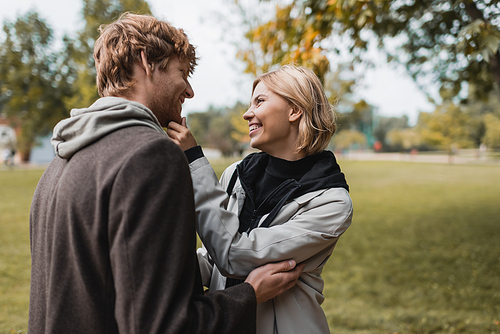 pleased couple in coats looking at each other while hugging in autumnal park during date