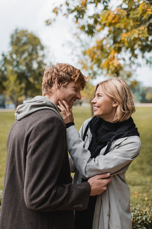 joyful couple in coats looking at each other while embracing in autumnal park during date