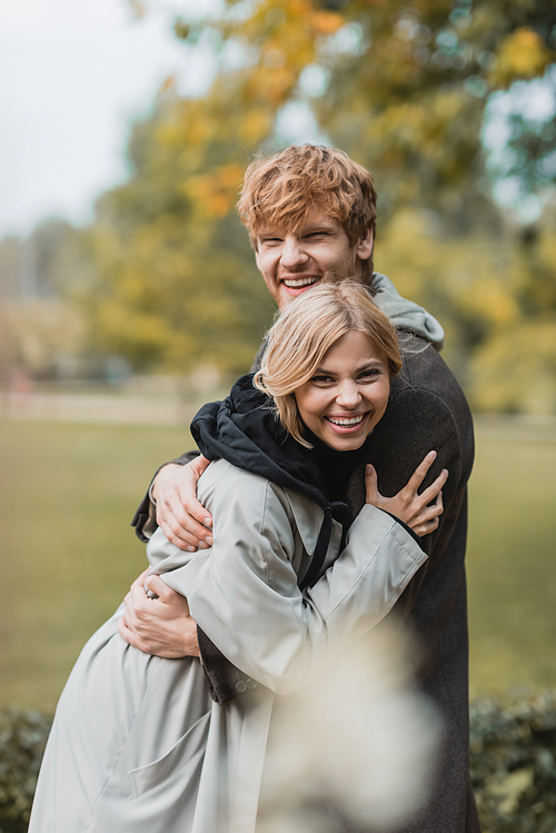 cheerful young man and woman in autumnal coats embracing each other and smiling in park