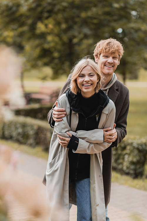 joyful redhead man embracing happy woman while smiling together during date in park