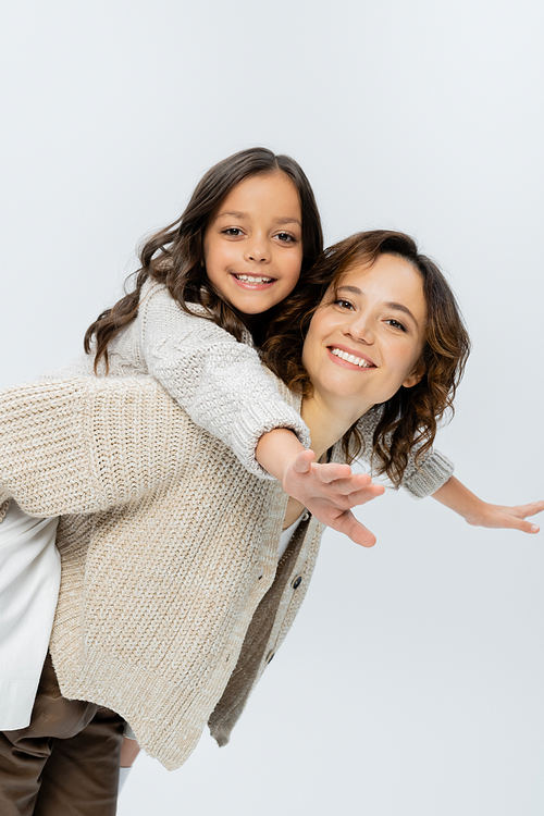 smiling woman playing with cheerful daughter while piggybacking her isolated on grey