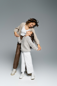 stylish mother and daughter holding hands and smiling at each other while dancing on grey background