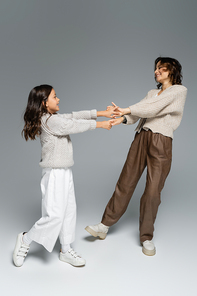 excited woman with daughter in autumn outfit dancing while holding hands on grey background
