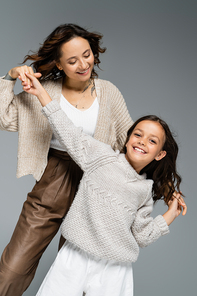 trendy and cheerful girl looking at camera while dancing with happy mom isolated on grey
