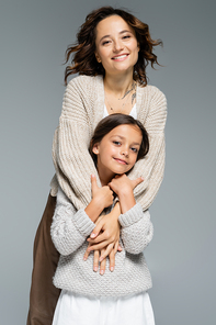 excited brunette woman in trendy knitwear hugging smiling daughter isolated on grey