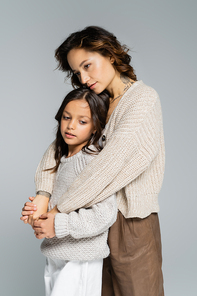 dreamy woman in trendy autumn clothes hugging daughter isolated on grey