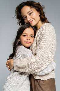 cheerful mom and daughter in cozy knitwear hugging and smiling at camera isolated on grey