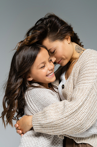 tattooed woman in warm clothes embracing excited daughter isolated on grey