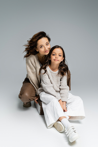 woman and child in trendy autumn outfit looking at camera while sitting on grey background