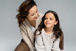 cheerful brunette woman in trendy and warm clothes looking at smiling daughter isolated on grey
