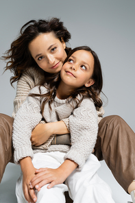stylish and happy woman looking at camera while hugging daughter on grey background