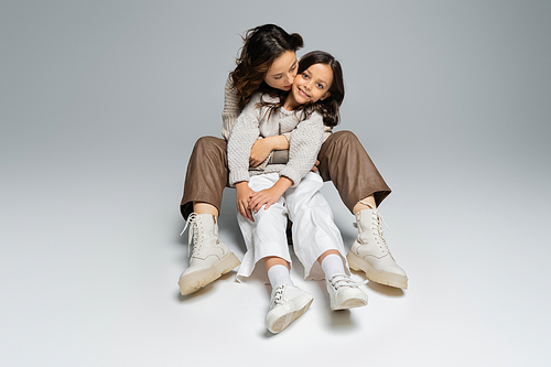 full length of stylish woman embracing daughter while sitting on grey background
