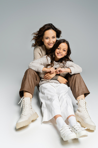 mother and child in trendy autumn clothes smiling at camera and sitting on grey