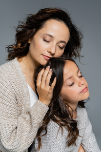 brunette woman with closed eyes embracing head of daughter isolated on grey