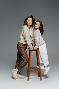 full length of mother and daughter in trendy autumn outfit posing near wooden stool on grey