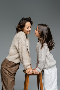 fashionable mother and daughter in warm knitwear looking at each other near wooden stool isolated on grey
