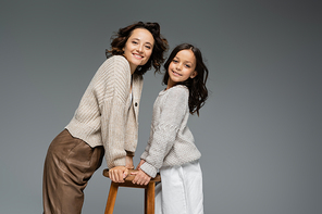 smiling mother and daughter in trendy autumn outfit posing near wooden stool isolated on grey
