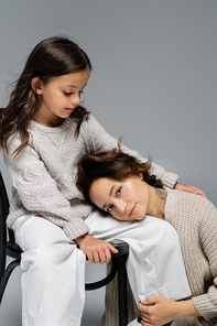 brunette woman looking at camera while leaning on trendy daughter sitting on chair on grey background