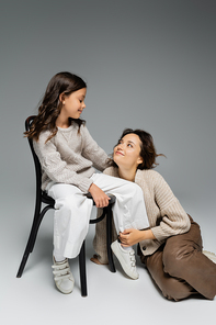 happy woman looking at daughter sitting on chair in trendy autumn outfit on grey background