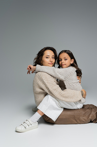 woman and child in warm knitwear hugging and looking at camera on grey background