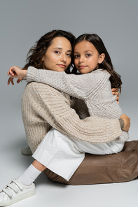 mother and daughter in warm clothes hugging and looking at camera on grey background