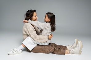 side view of brunette mother and daughter in warm clothes smiling at each other on grey background