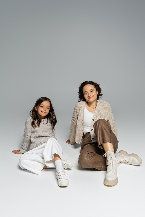 brunette mother and daughter in knitwear and pants sitting on grey background