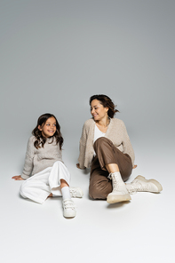 smiling woman looking at trendy daughter while sitting on grey background