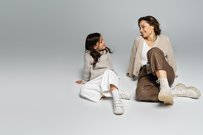 full length of fashionable mom and daughter smiling at each other on grey background
