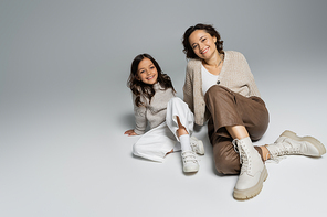 girl and mother in trendy and warm clothes sitting and smiling at camera on grey background