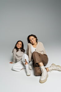 full length of mother and daughter in knitwear and pants sitting on grey background