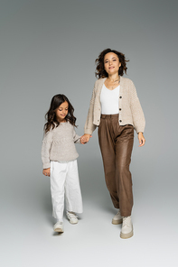 trendy woman smiling at camera while holding hands with daughter and walking on grey background