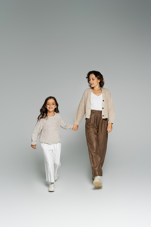 full length of mom and daughter in warm knitwear and pants walking while holding hands on grey background